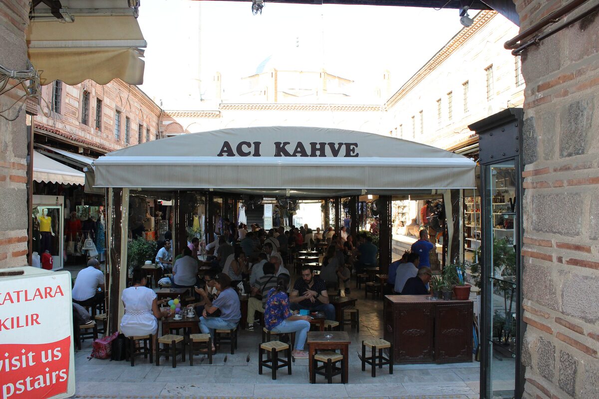 Café tables under a white awning in the courtyard of Kızlarağası Han, surrounded by the han's striped stone and brick arcades