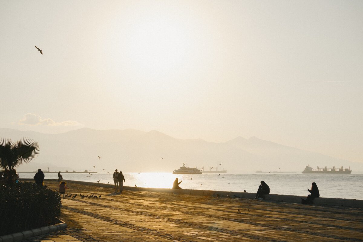 People sitting and walking on the Kordon waterfront at sunset, with ships on the gulf and the mountains behind them