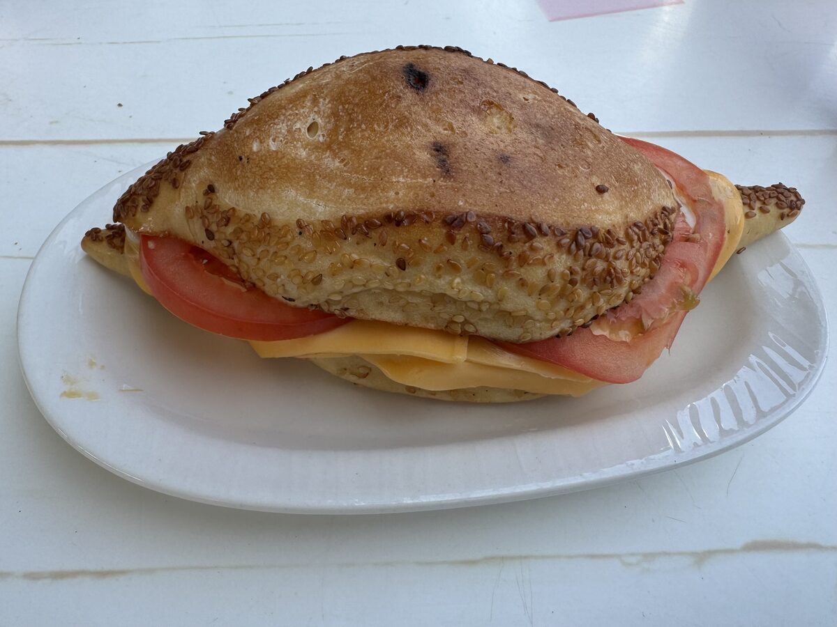 A kumru sandwich on a white plate, a sesame-covered roll filled with melted cheese, sausage and tomato