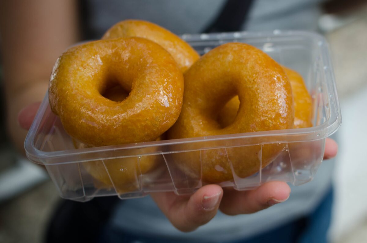 A hand holding a plastic box of golden, ring-shaped lokma soaked in syrup