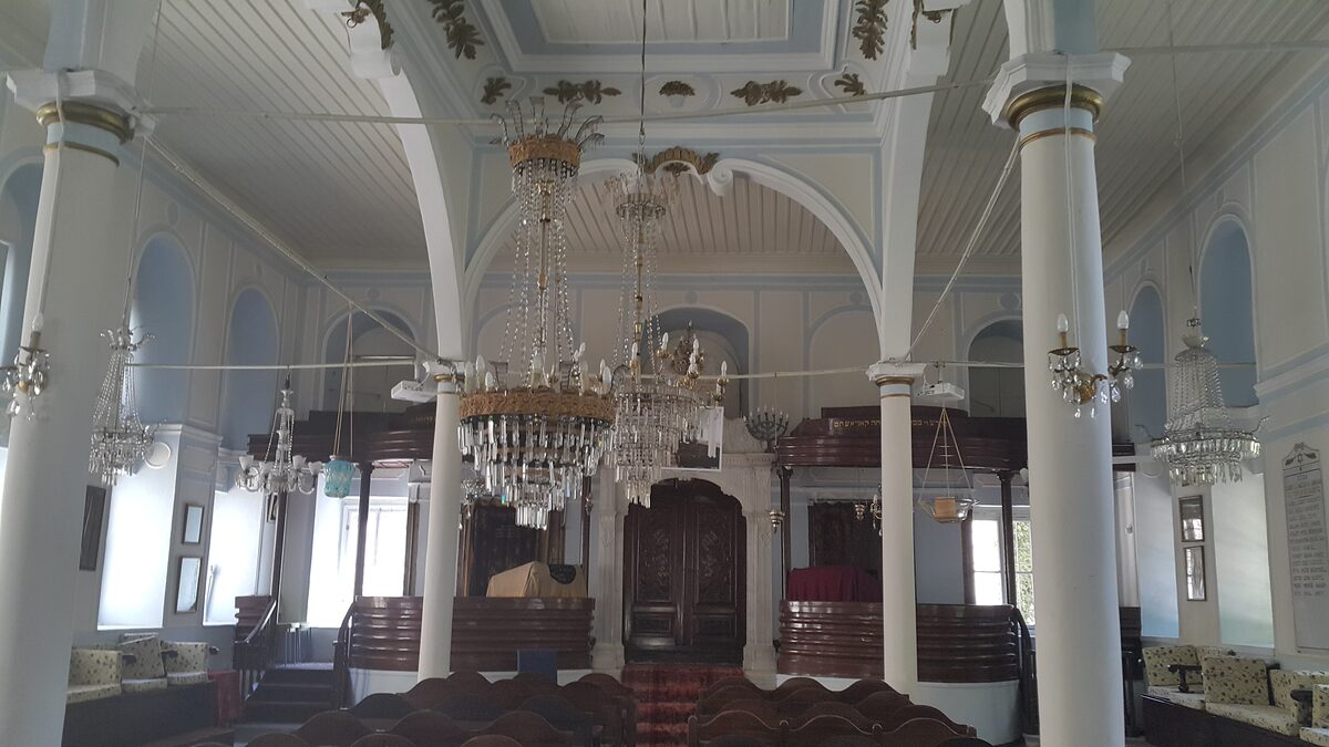 The white interior of the Sinyora Giveret synagogue, with slim columns, crystal chandeliers and wooden pews facing the carved ark
