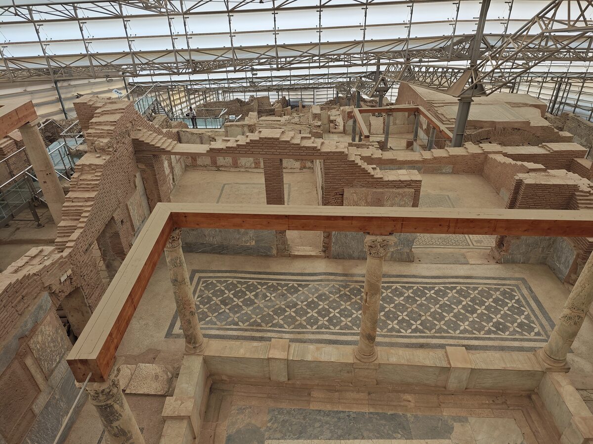 Excavated Roman houses under a modern protective roof at Ephesus, seen from a walkway, with mosaic floors and painted walls