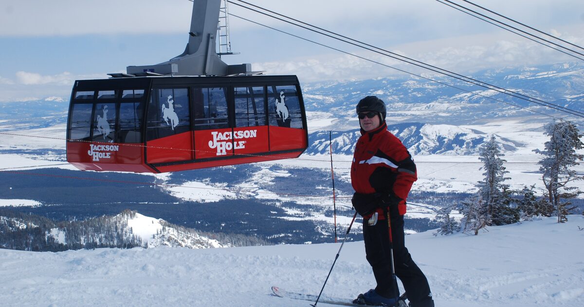 A skier in a red jacket on the snow at Jackson Hole Mountain Resort beside a red Aerial Tram car, with the winter valley below