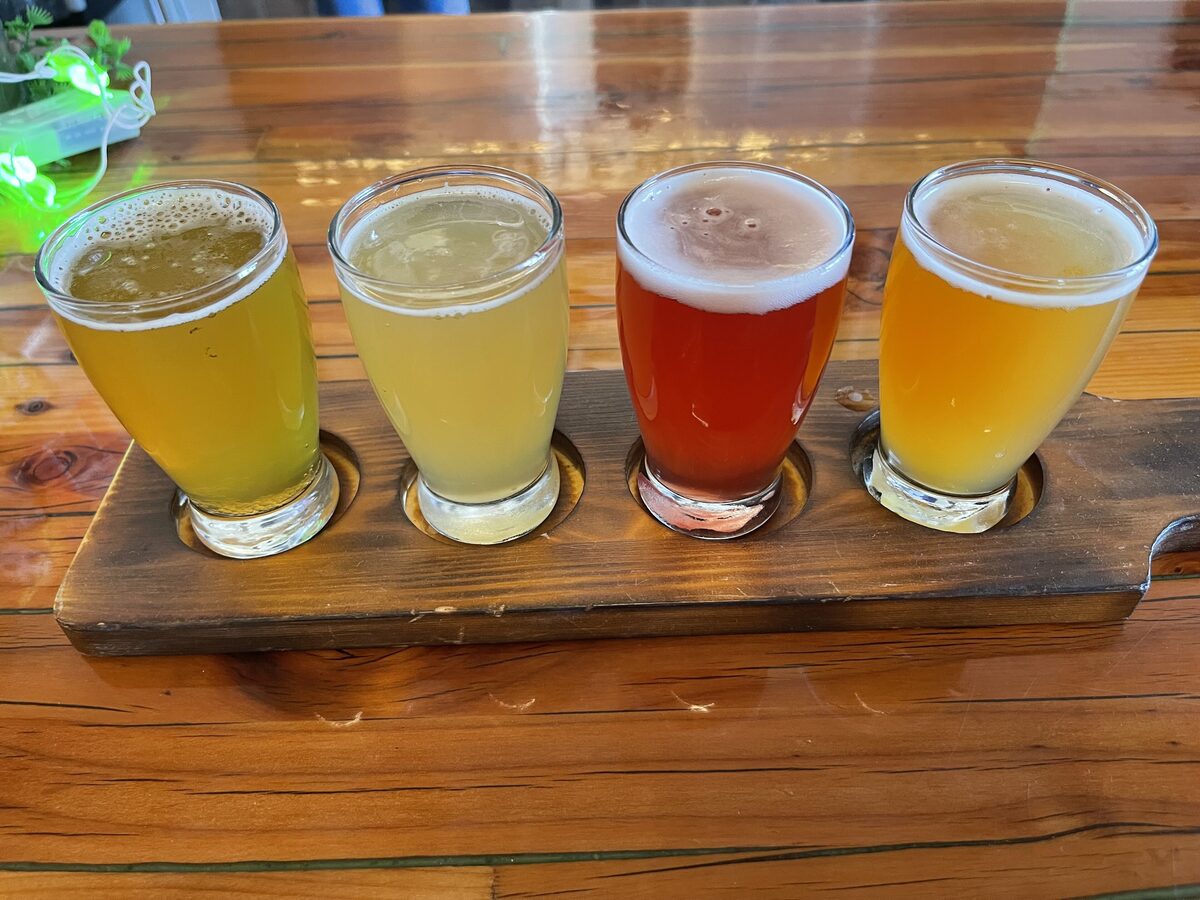 A wooden paddle holding a flight of four craft beers in small glasses, from pale gold to red amber