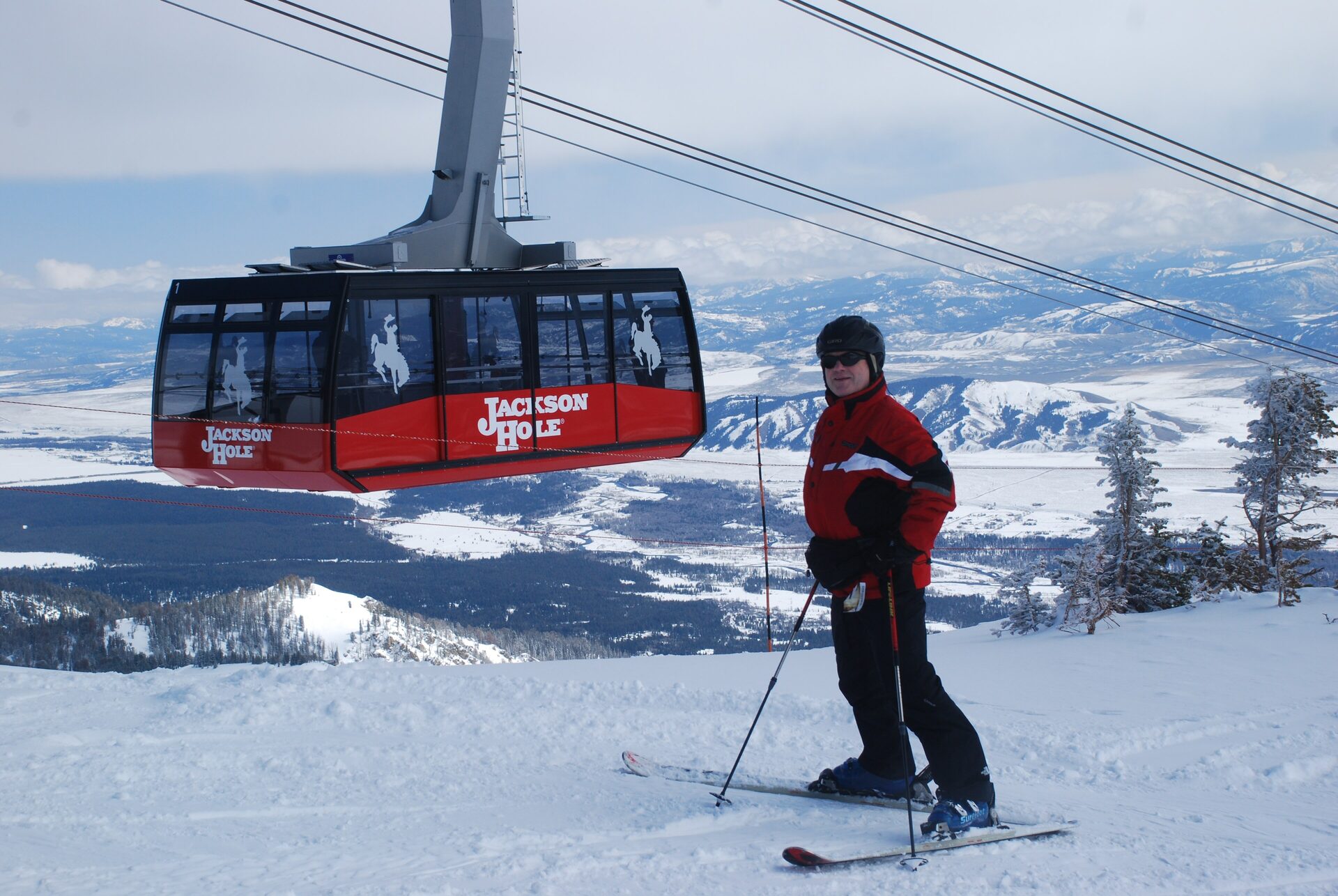 A skier in a red jacket on the snow at Jackson Hole Mountain Resort beside a red Aerial Tram car, with the winter valley below