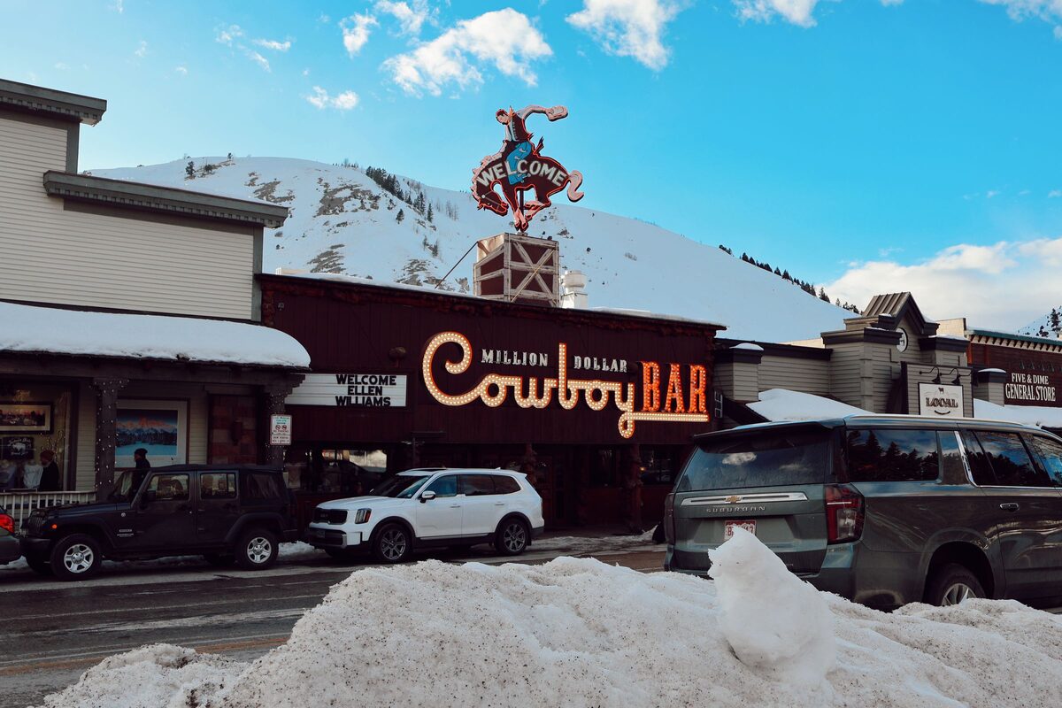 The Million Dollar Cowboy Bar in Jackson on a snowy day, its neon sign and a cowboy figure above the roof, with a snow-covered hill behind