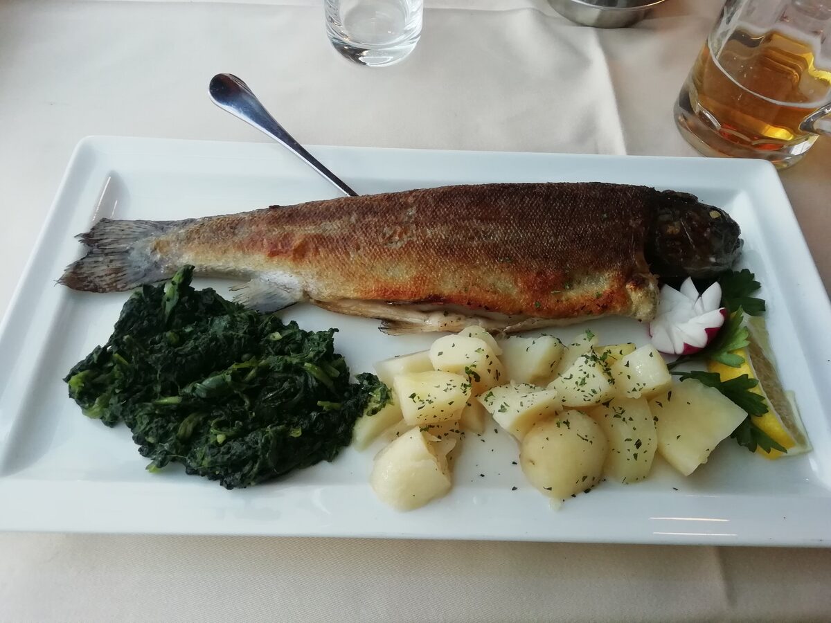 A whole pan-fried trout on a white plate with spinach and boiled potatoes