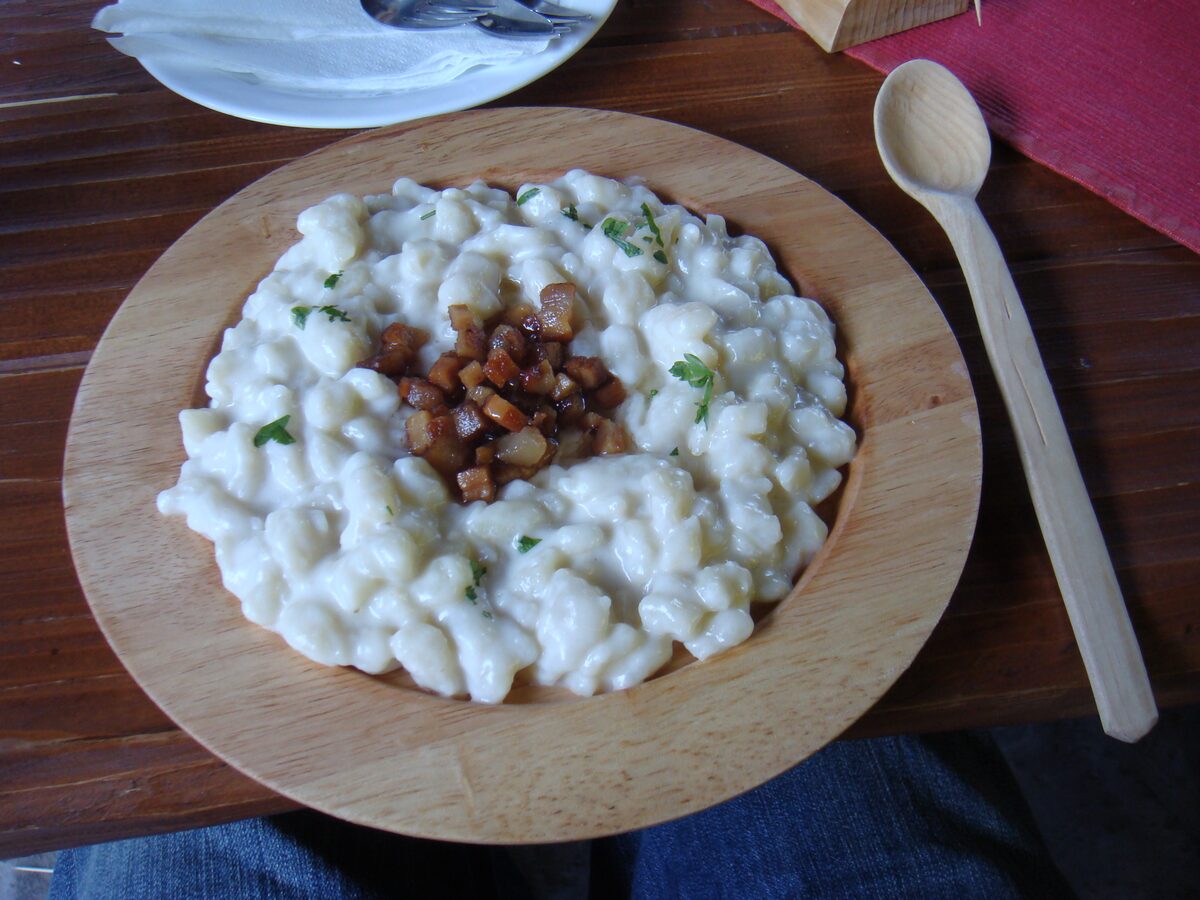 Small white potato dumplings in a creamy sauce on a wooden plate, with cubes of fried bacon in the middle and a wooden spoon beside it