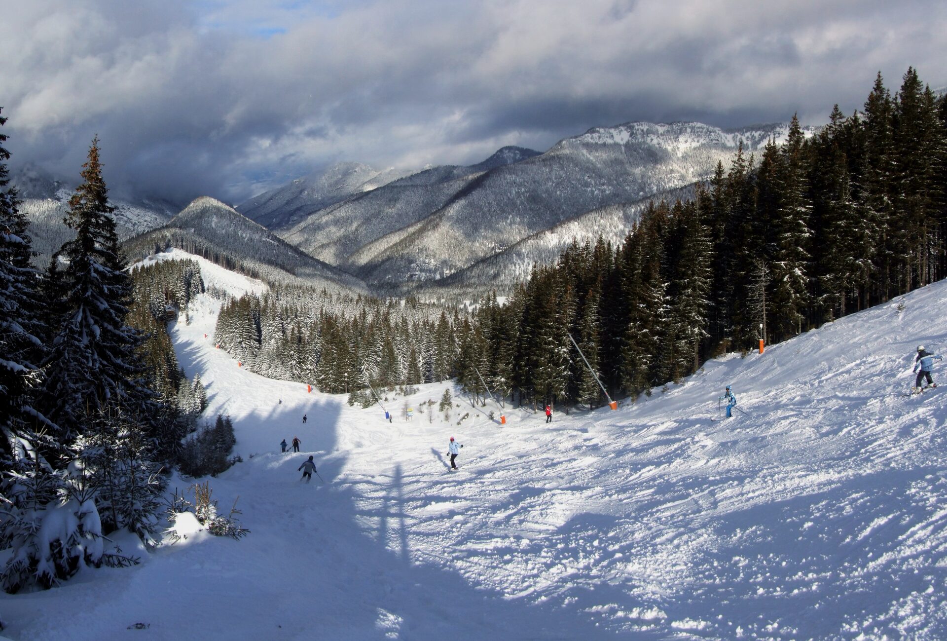 Skiers on a groomed run in Jasná with snowy, forested mountains above