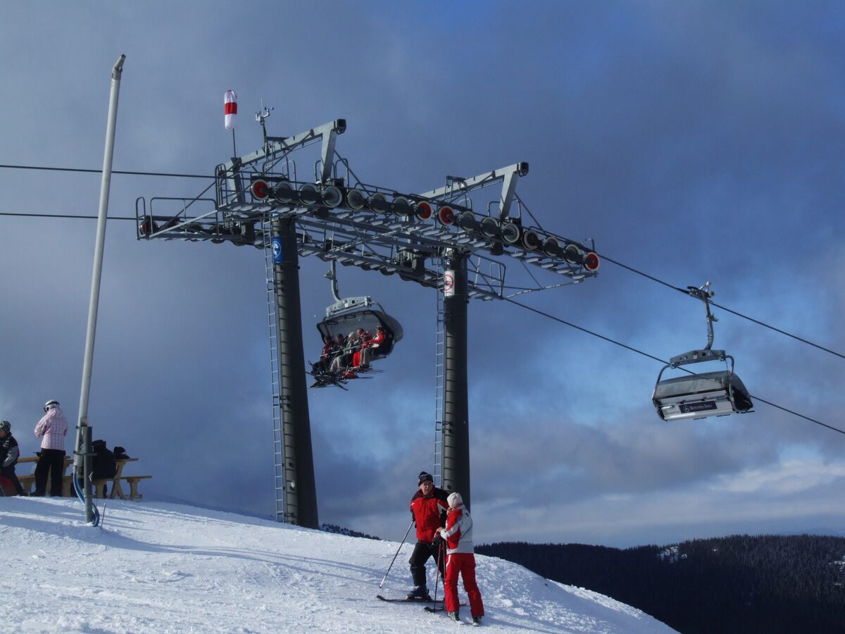 A chairlift station in snow with cabins overhead and two people in red ski clothes beside it