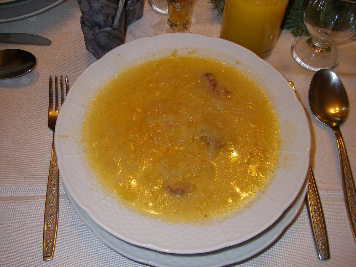 A white bowl of yellow sauerkraut soup with pieces of sausage, set on a table with a fork and spoon