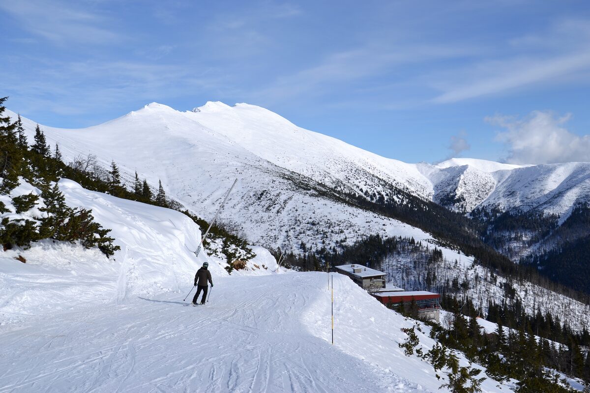 A skier on a snowy Jasná slope with the Ďumbier ridge and a lodge in the distance