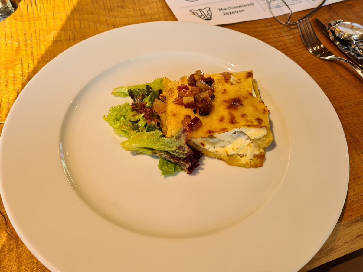 A folded potato flatbread filled with white cheese, topped with cubes of bacon, on a large white plate with a little salad