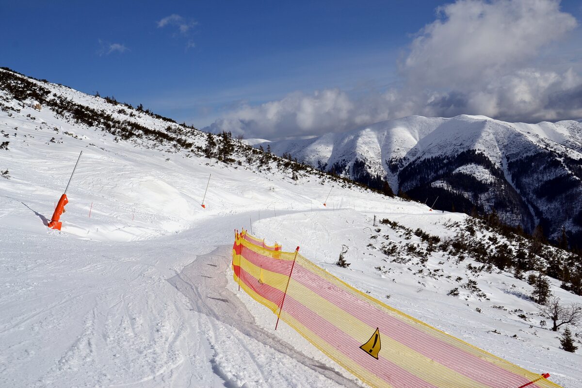 A wide snowy piste with orange safety netting along its edge and mountains behind