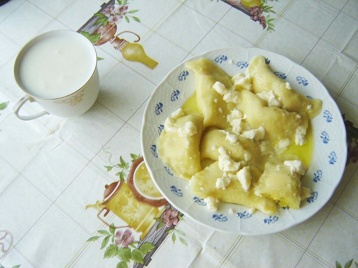 Filled dumplings topped with crumbled white cheese on a plate with a blue flower pattern, next to a cup of white drink