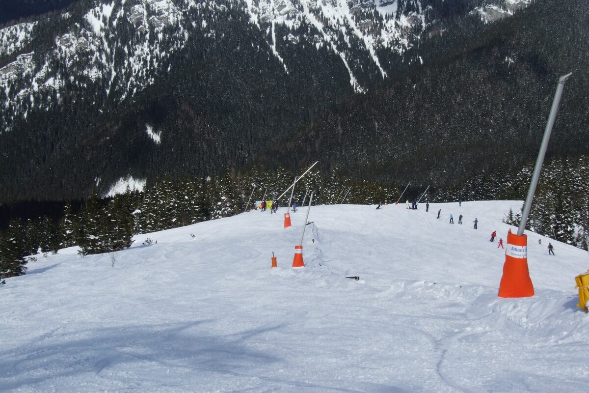 A red intermediate piste with snow-making poles and spruce forest along its edge