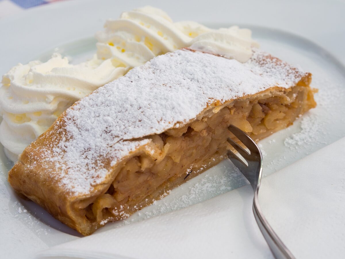 A slice of apple strudel dusted with icing sugar, with whipped cream and a fork