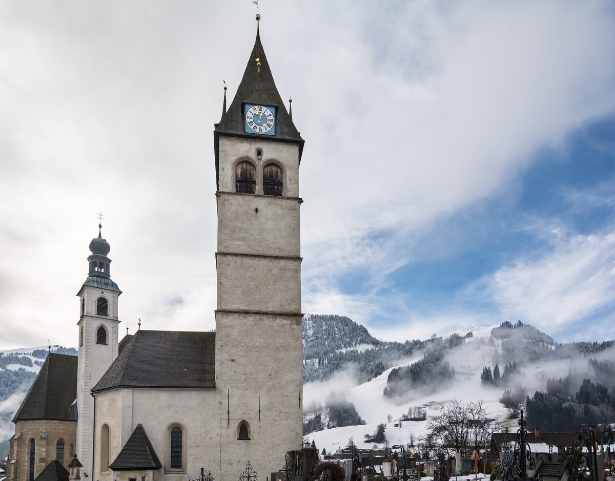 The tower of St. Andrew's parish church in Kitzbühel, with a smaller onion-domed spire beside it and misty slopes behind