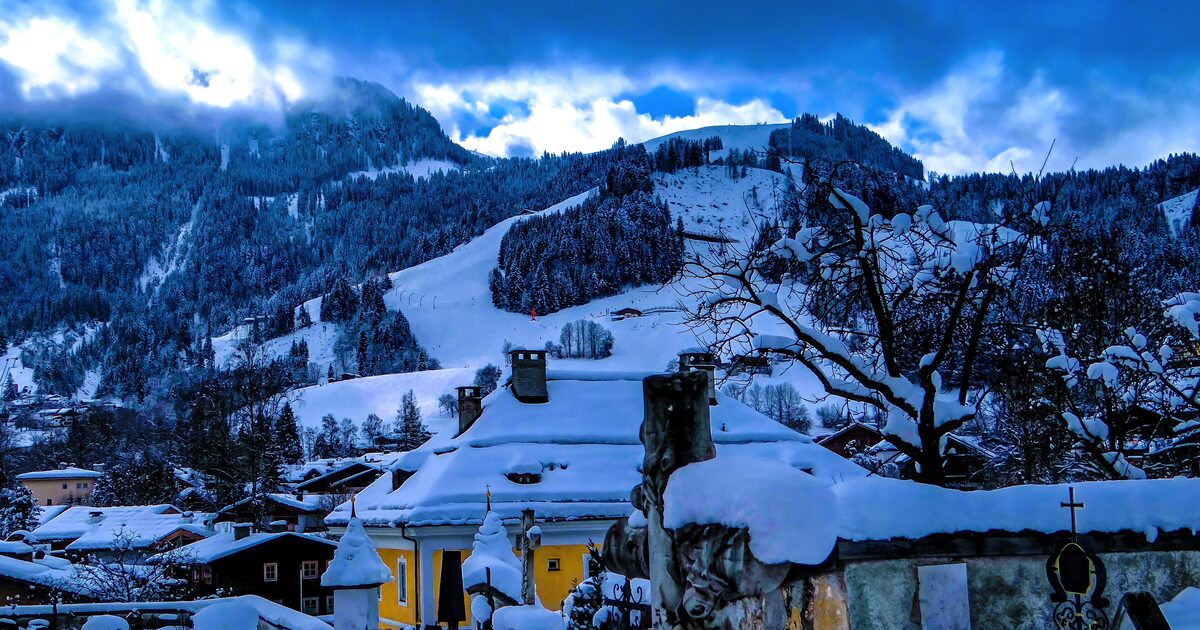 Snow-covered Hahnenkamm above the rooftops and churchyard of Kitzbühel, with a ski slope running down under dark winter clouds