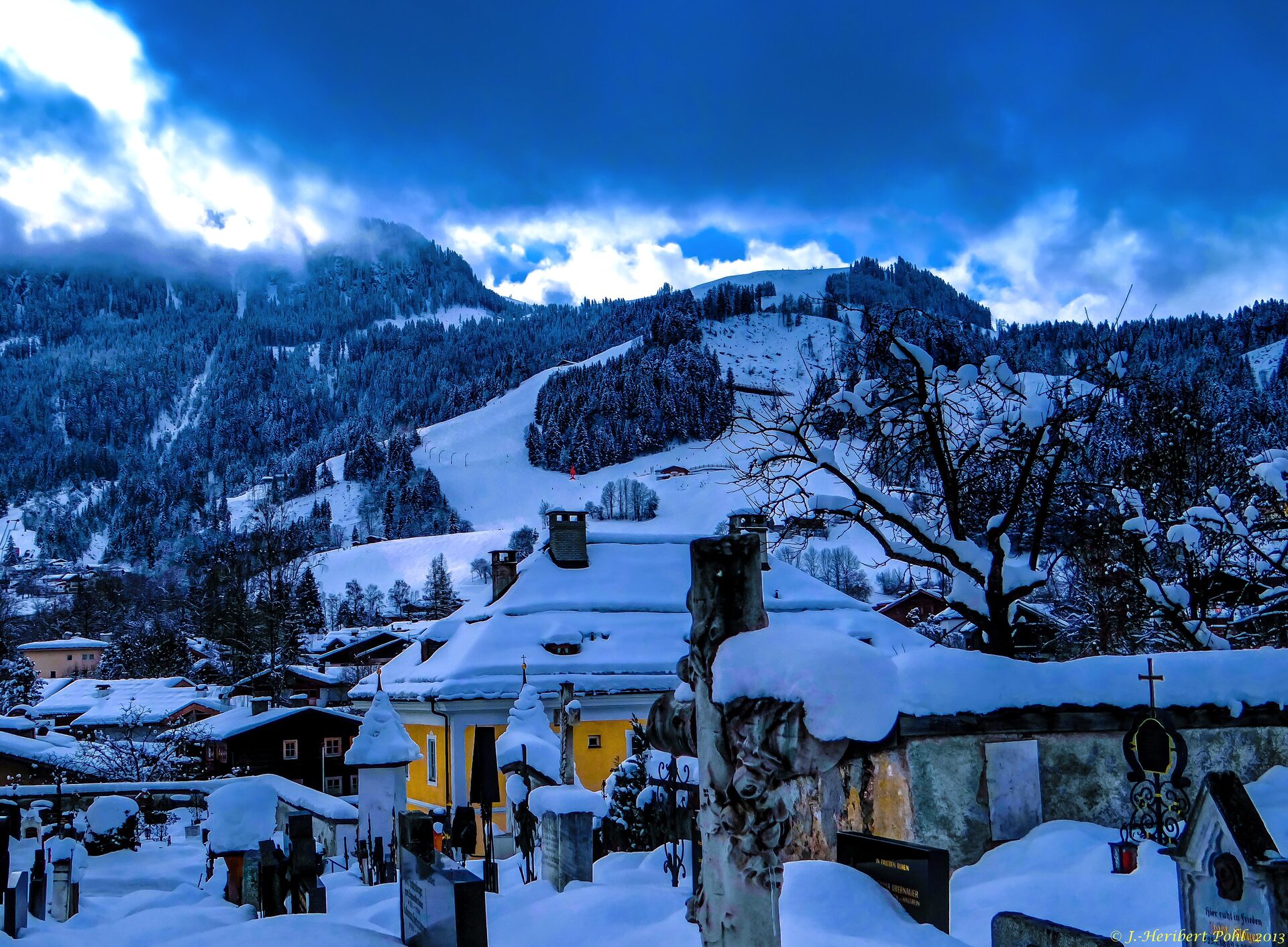 Snow-covered Hahnenkamm above the rooftops and churchyard of Kitzbühel, with a ski slope running down under dark winter clouds