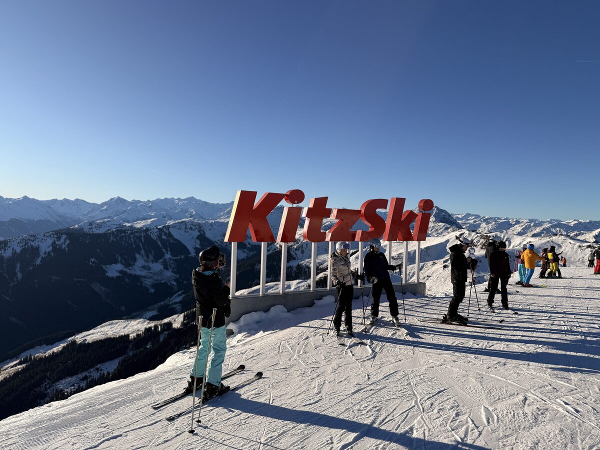 Skiers stopping by the red KitzSki lettering on a sunny, groomed ridge, with snowy peaks behind