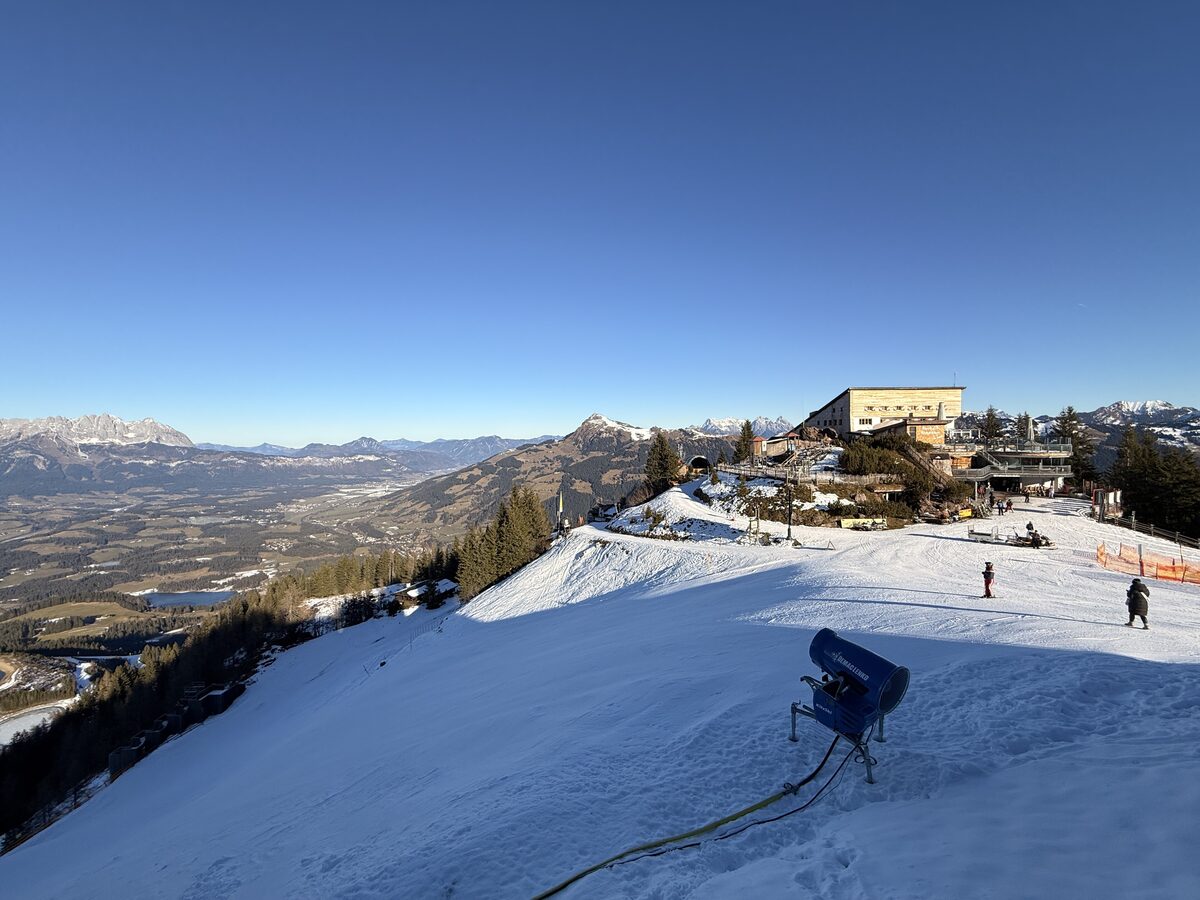 Snowmaking gun on a groomed slope next to the station building at the top of the Hahnenkamm, with the valley and the Kaiser mountains far below