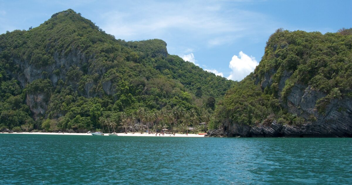 Limestone cliffs and forest above a small white sand beach with palms, seen from the sea in Ang Thong Marine Park