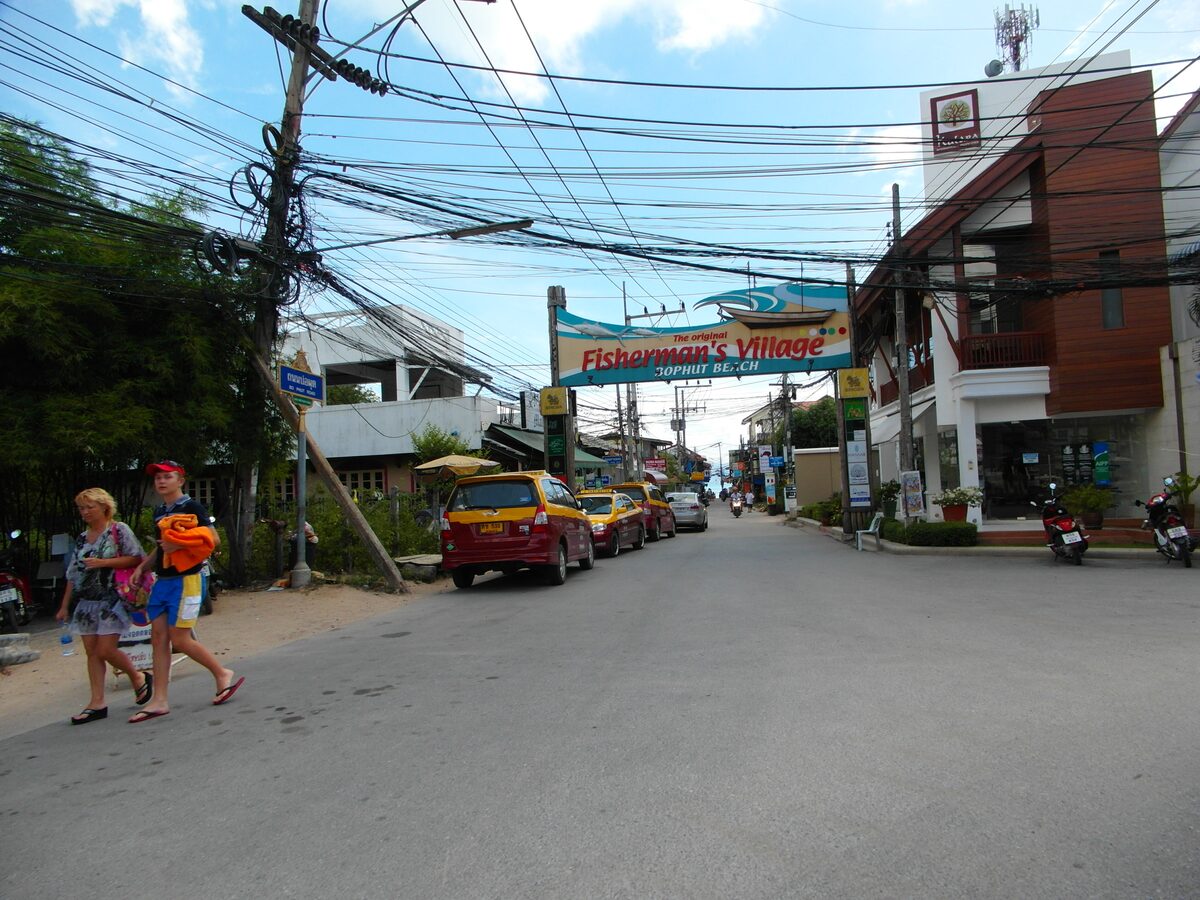 The Fisherman's Village sign over the road in Bophut, with cars and shops along the street