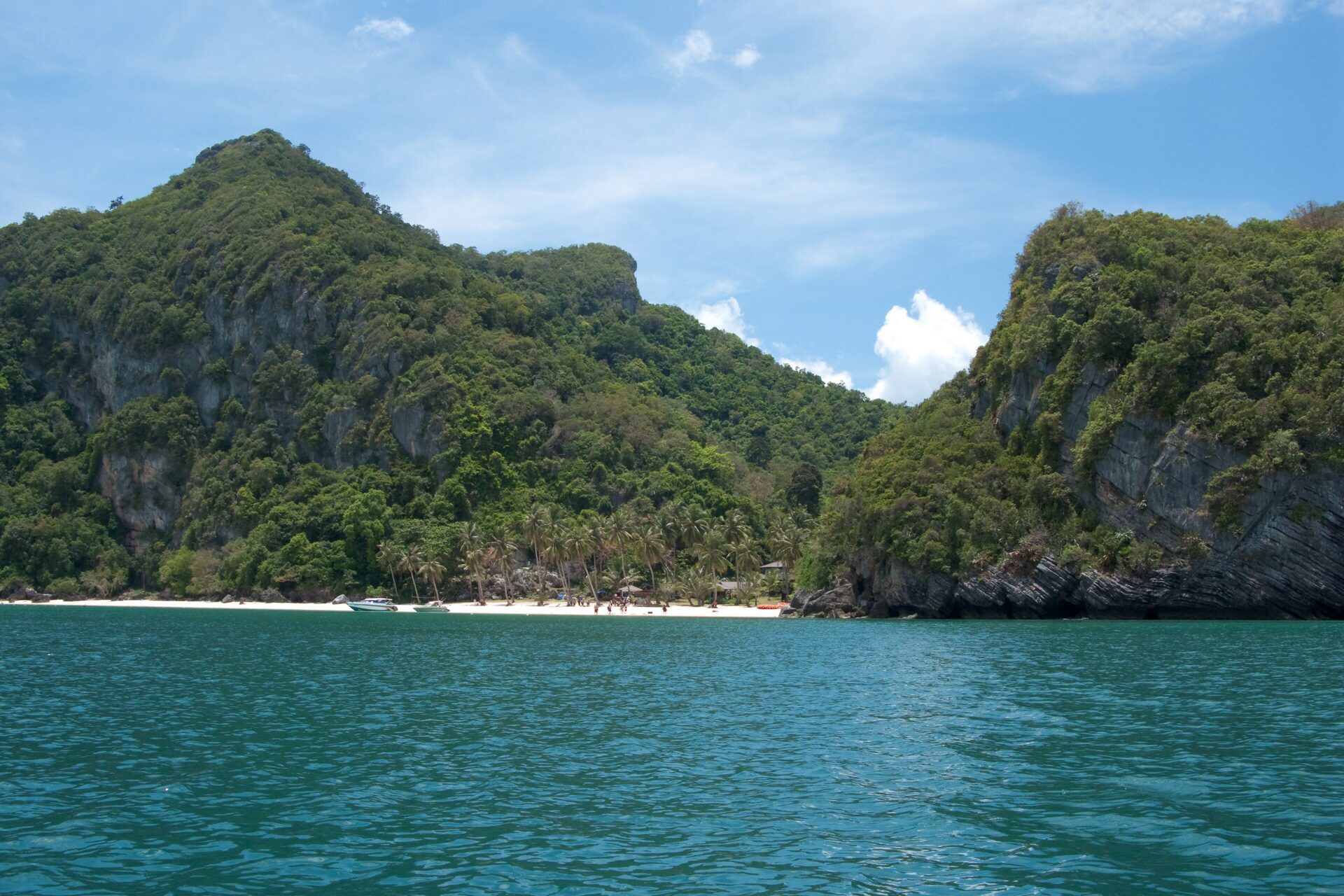 Limestone cliffs and forest above a small white sand beach with palms, seen from the sea in Ang Thong Marine Park
