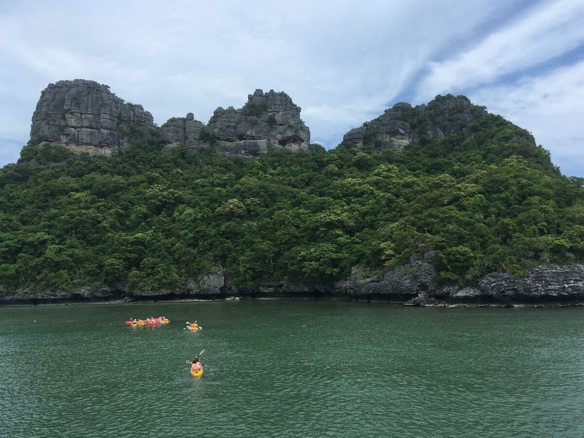 A small group of sea kayakers paddling in green water below the limestone peaks of Ko Mae Ko in Ang Thong