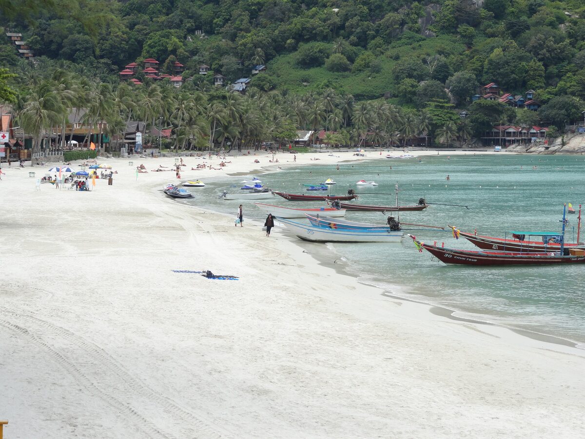 White sand and long-tail boats along Haad Rin Sunrise Beach on Koh Phangan, with forested hills behind