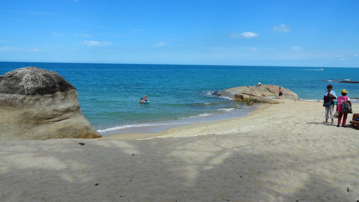Granite boulders on the sand at Lamai Beach, with bright turquoise water, a swimmer and two people standing on the right