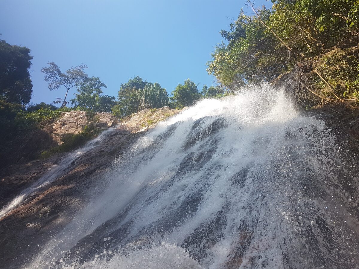 Water sliding down a smooth rock face at Na Muang Waterfall, with forest and blue sky above