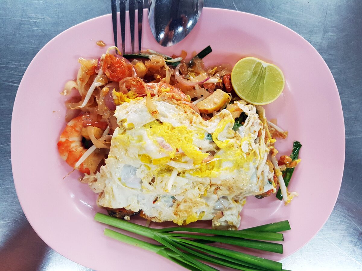 Pad Thai with shrimp under a folded egg omelette on a pink plate, with a lime wedge and garlic chives