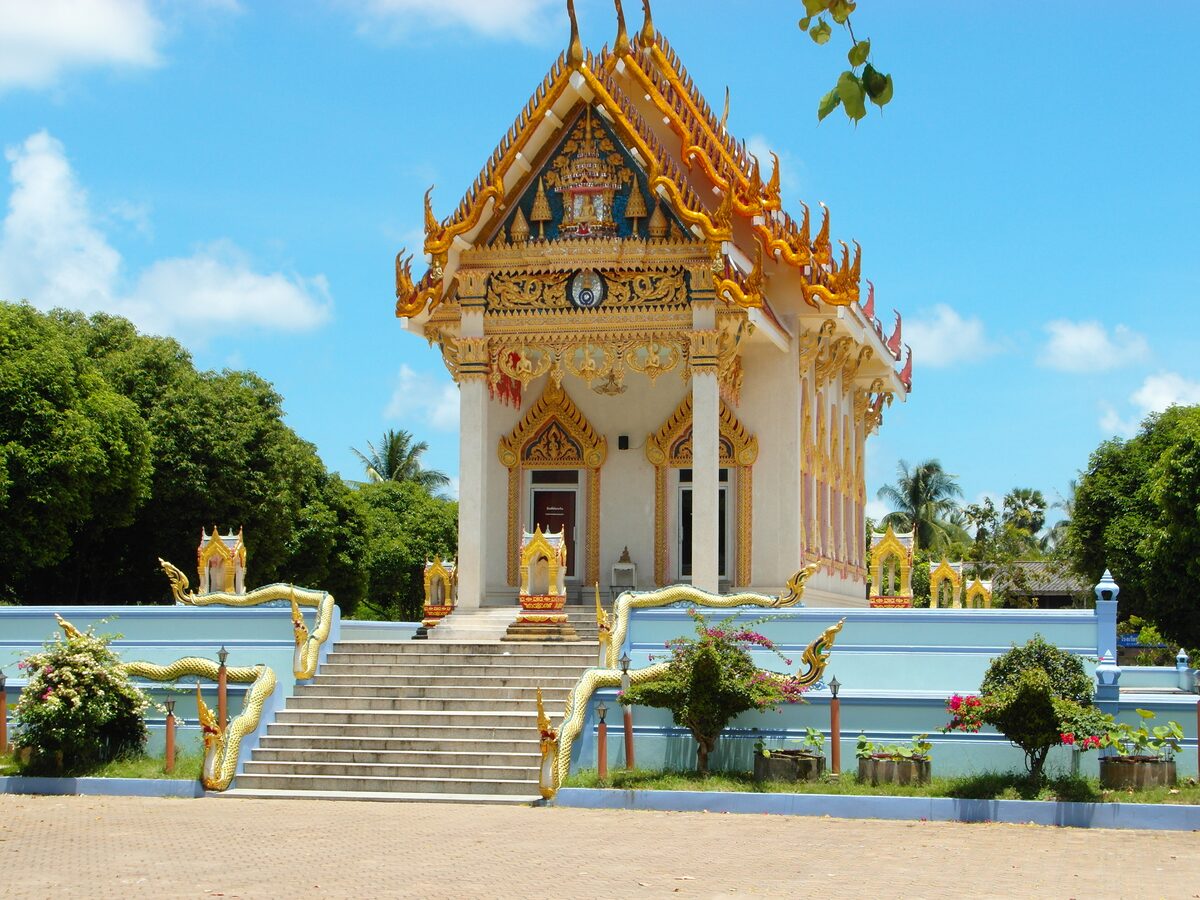 The white and gold temple hall of Wat Khunaram at the top of a stair guarded by naga railings, under a blue sky