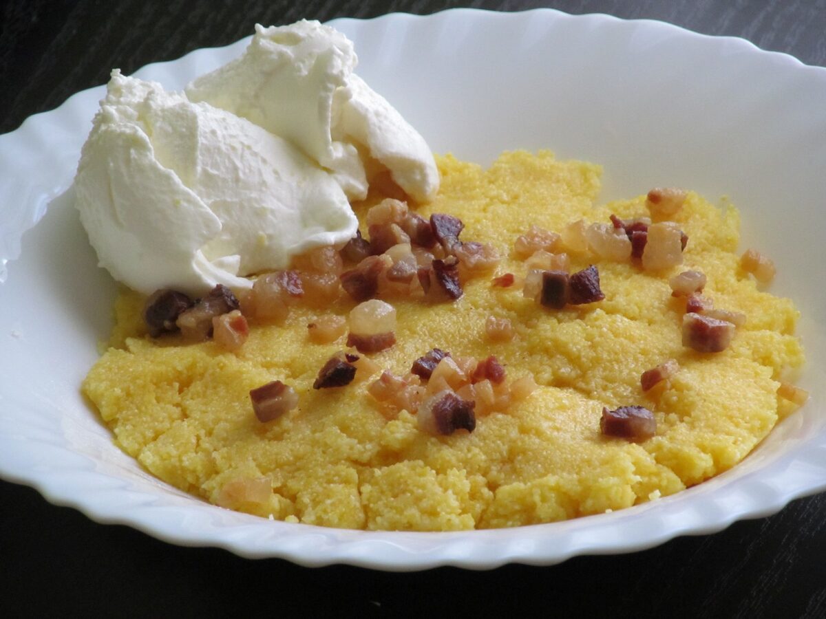 Cicvara on a white plate: yellow cornmeal porridge topped with a spoon of white cream and crisp bits of smoked meat