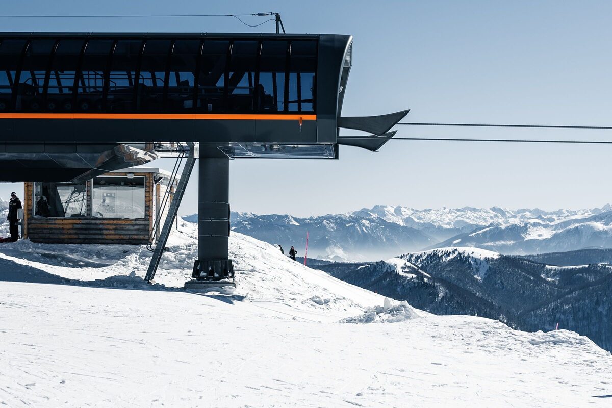 A chairlift station on a snowy ridge above Kolašin, with a ski lodge below it and snow-covered mountains behind under a clear blue sky