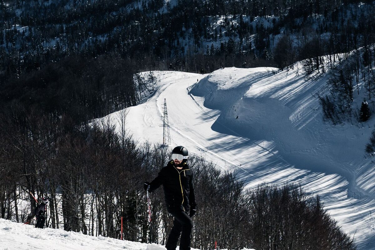 A skier in a black jacket and goggles standing on a forest-edged groomed slope near Kolašin, with a wide white run behind