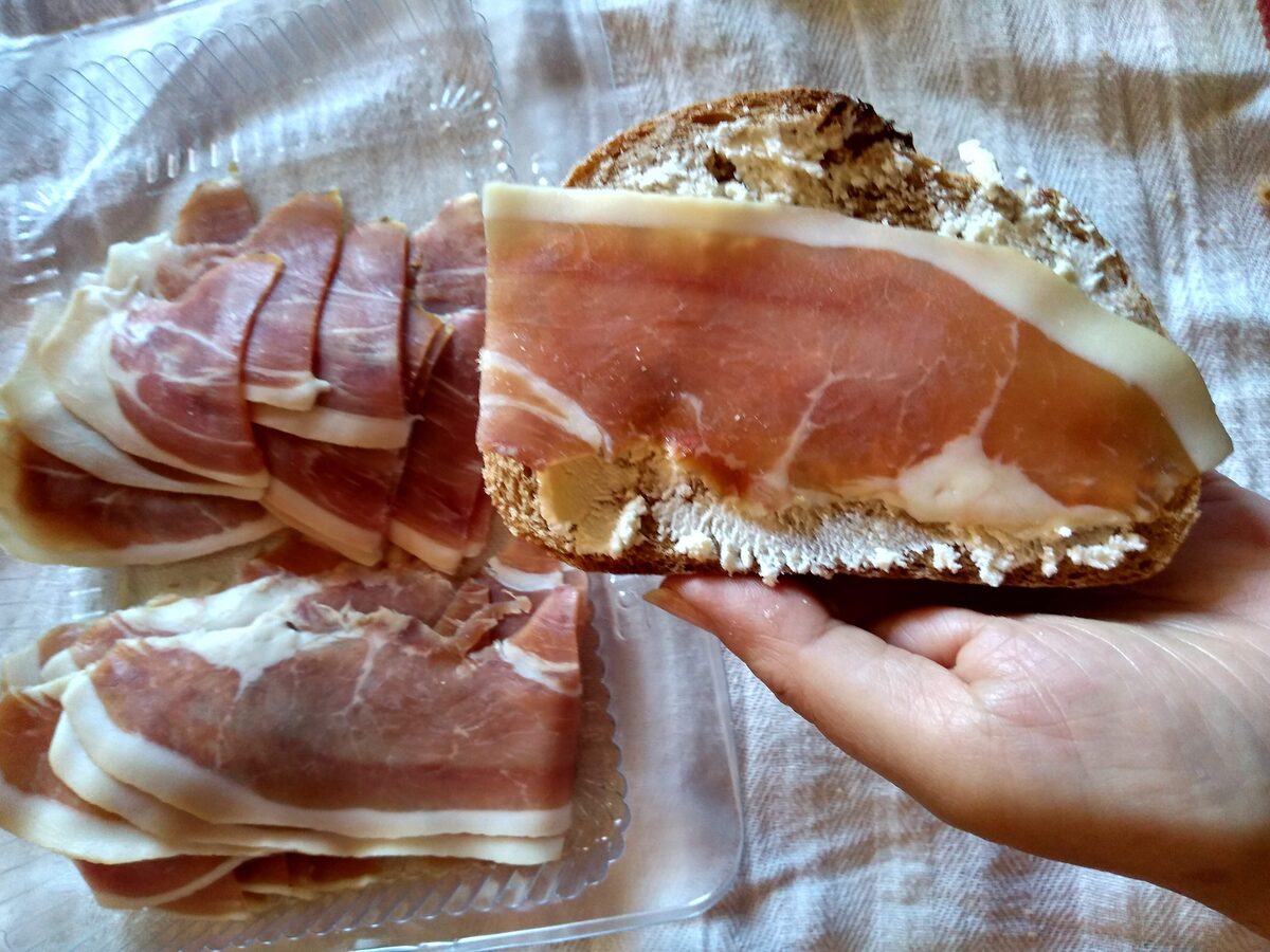 Slices of Njeguški pršut on a piece of rustic bread held in a hand, with white kajmak on the bread