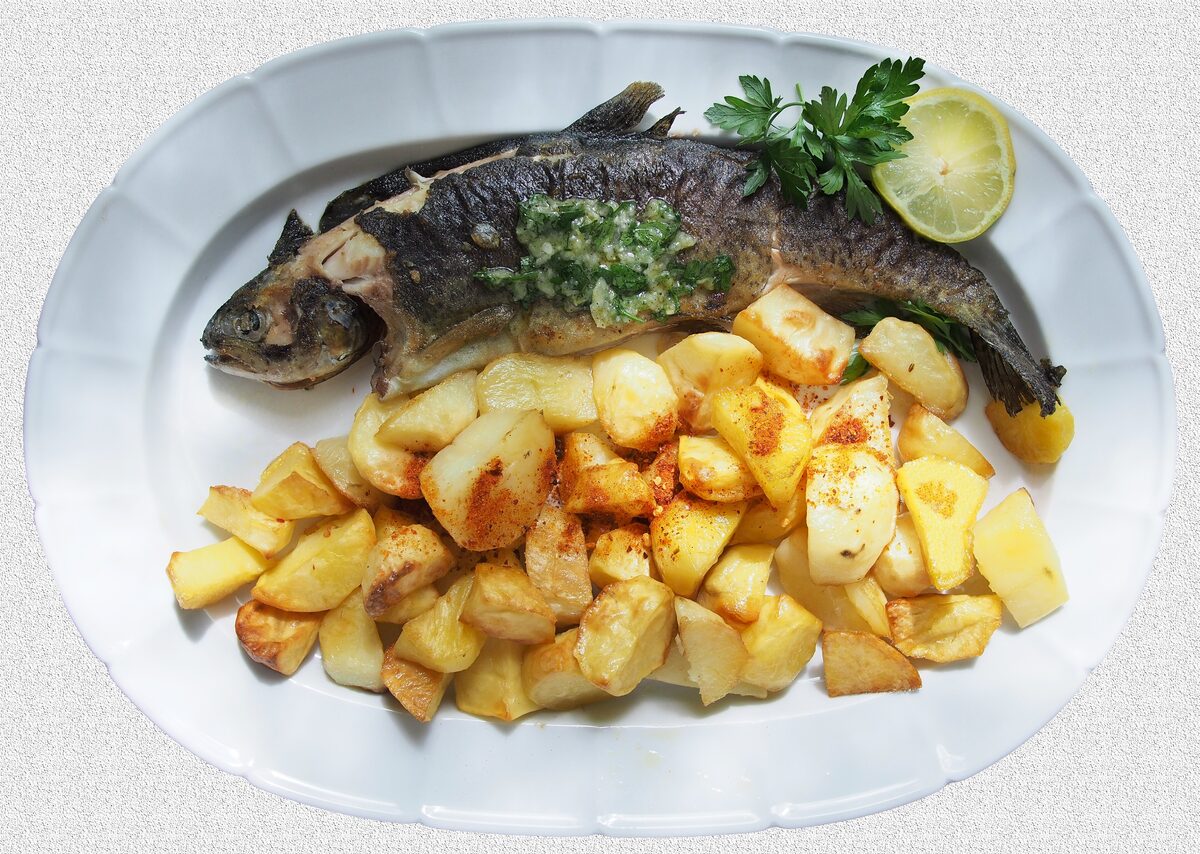 A whole baked trout with herb sauce and a lemon slice on a plate of roasted potatoes