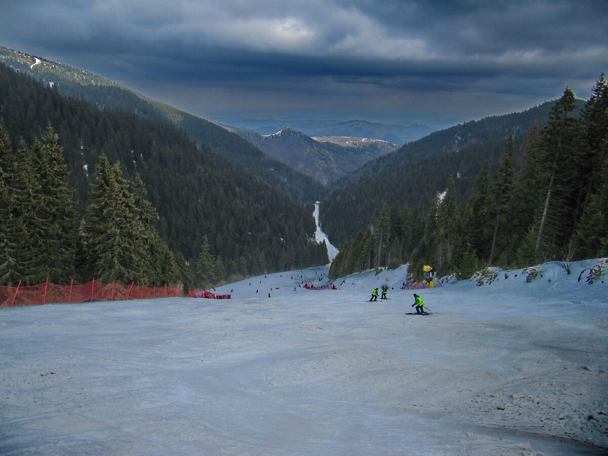 A wide groomed run with a few skiers dropping into a forested valley on Kopaonik, under a dark, stormy sky