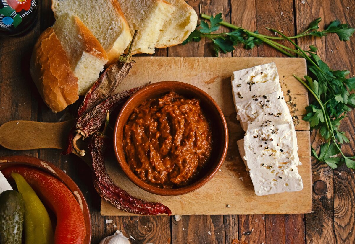 A bowl of ajvar next to slices of white cheese, bread, dried peppers and parsley on a wooden board