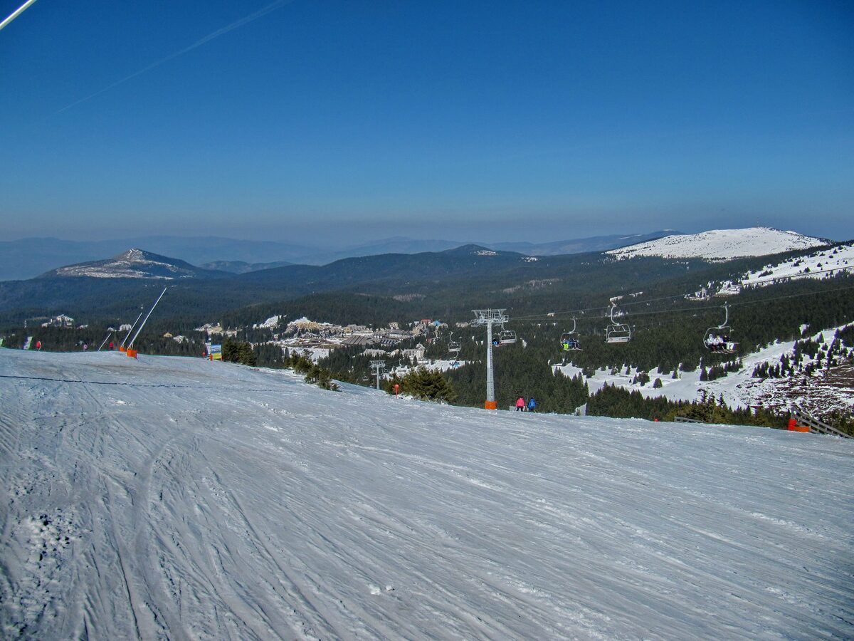 A chairlift over a groomed run on Kopaonik, with the resort buildings and distant peaks below under a blue sky