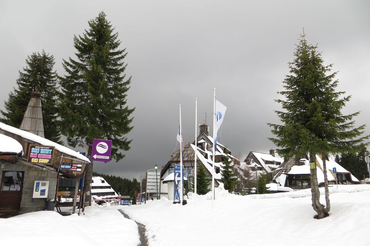 The resort centre in snow, with a ski rental shop, ski school sign and wooden-roofed buildings under a dark grey sky