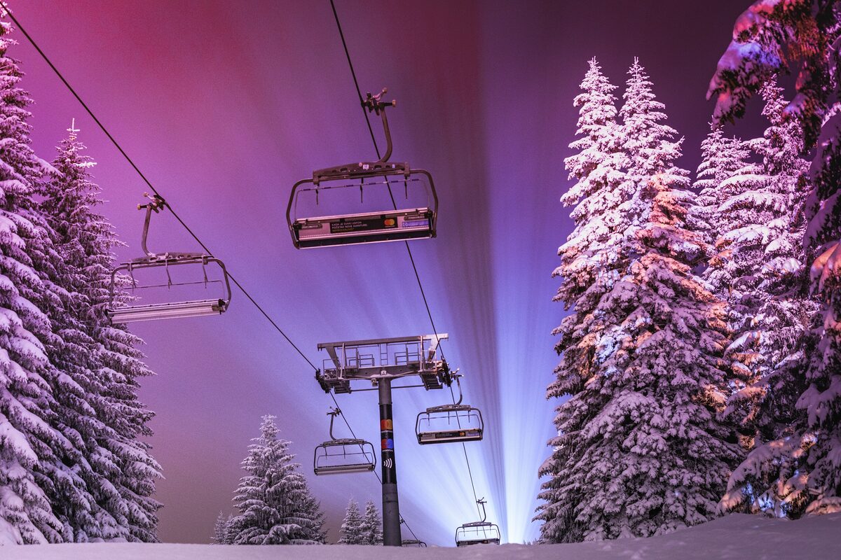 A chairlift lit up at night between snow-laden spruce trees, with the floodlights glowing purple and blue