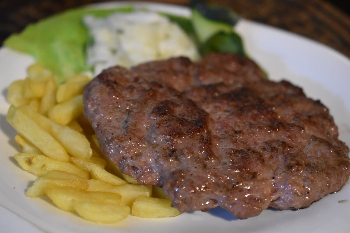 A grilled pljeskavica patty with fries and a creamy side salad on a white plate