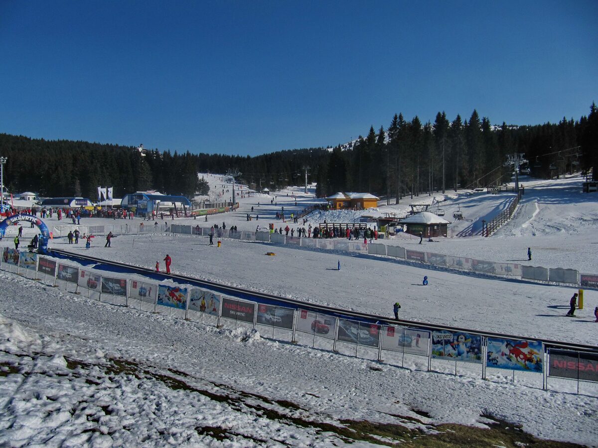 A fenced beginner area at the foot of a chairlift, with ski-school groups, a moving belt and a mountain hut