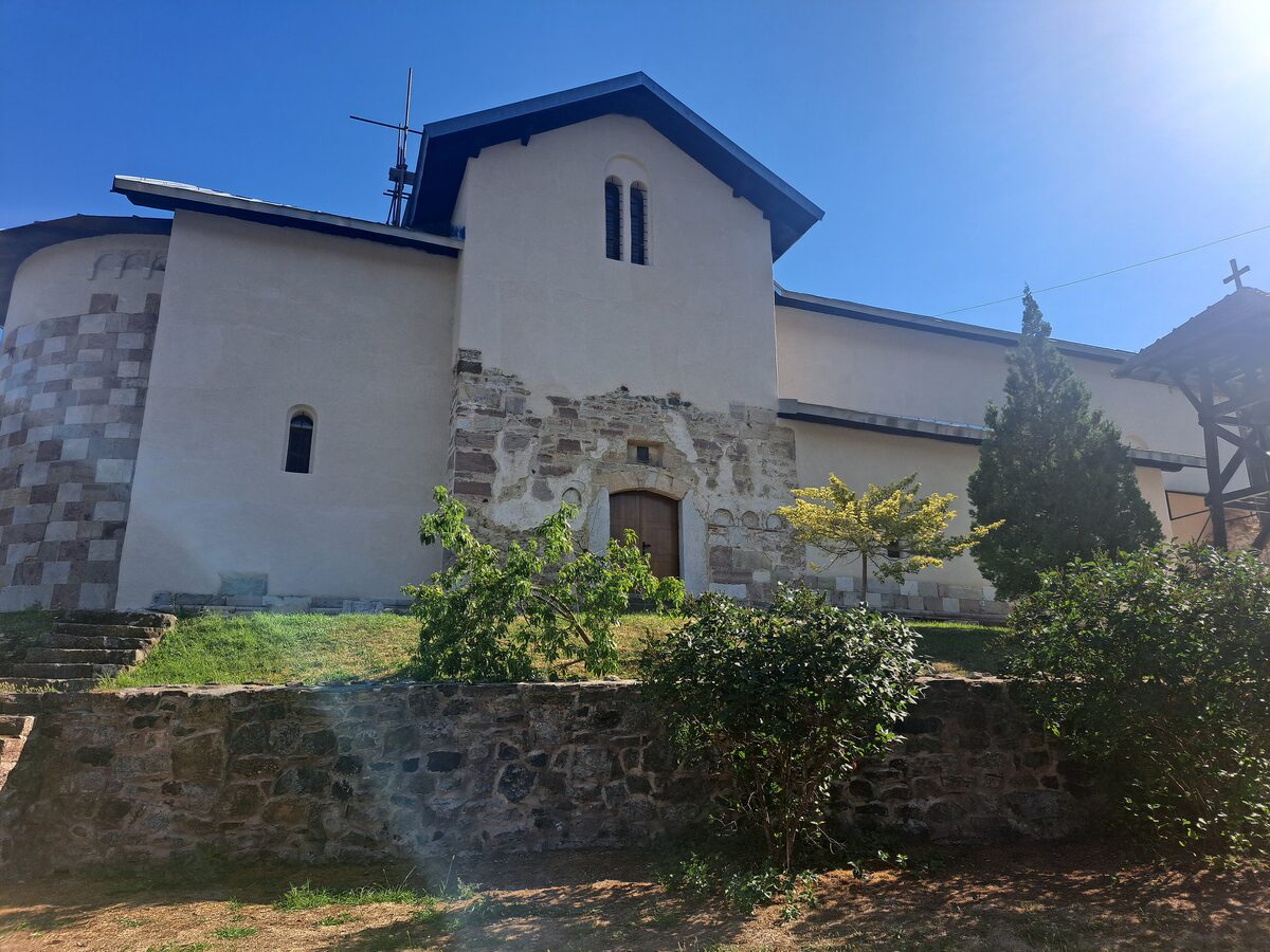 The church of Banjska monastery in summer sun, its surviving medieval portal and banded stone apse set into newer white plastered walls, above a low stone terrace wall.