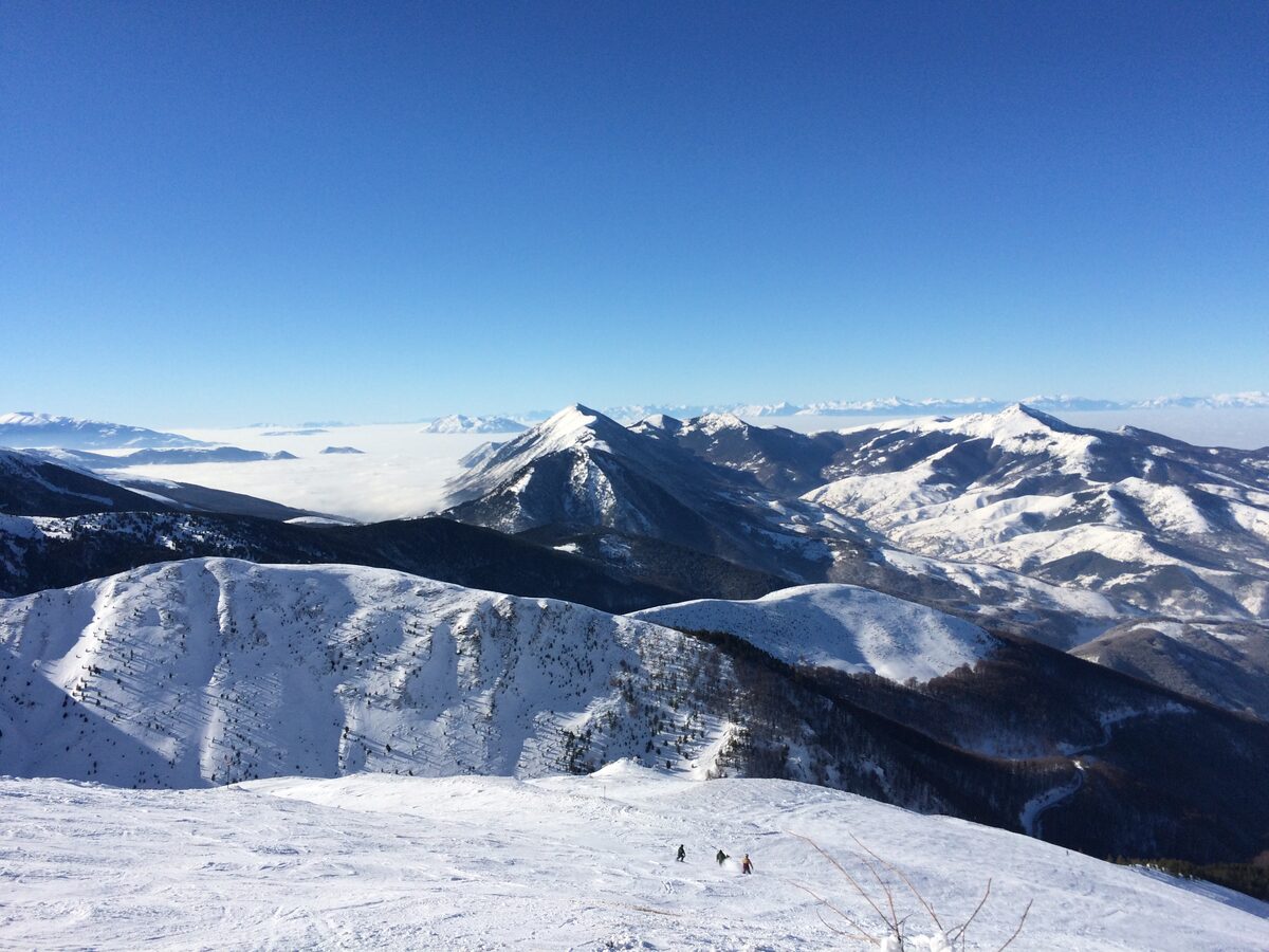 Snow-covered Šar mountain peaks seen from the top of the Brezovica ski slopes, with a few skiers below and a sea of cloud filling the valleys.
