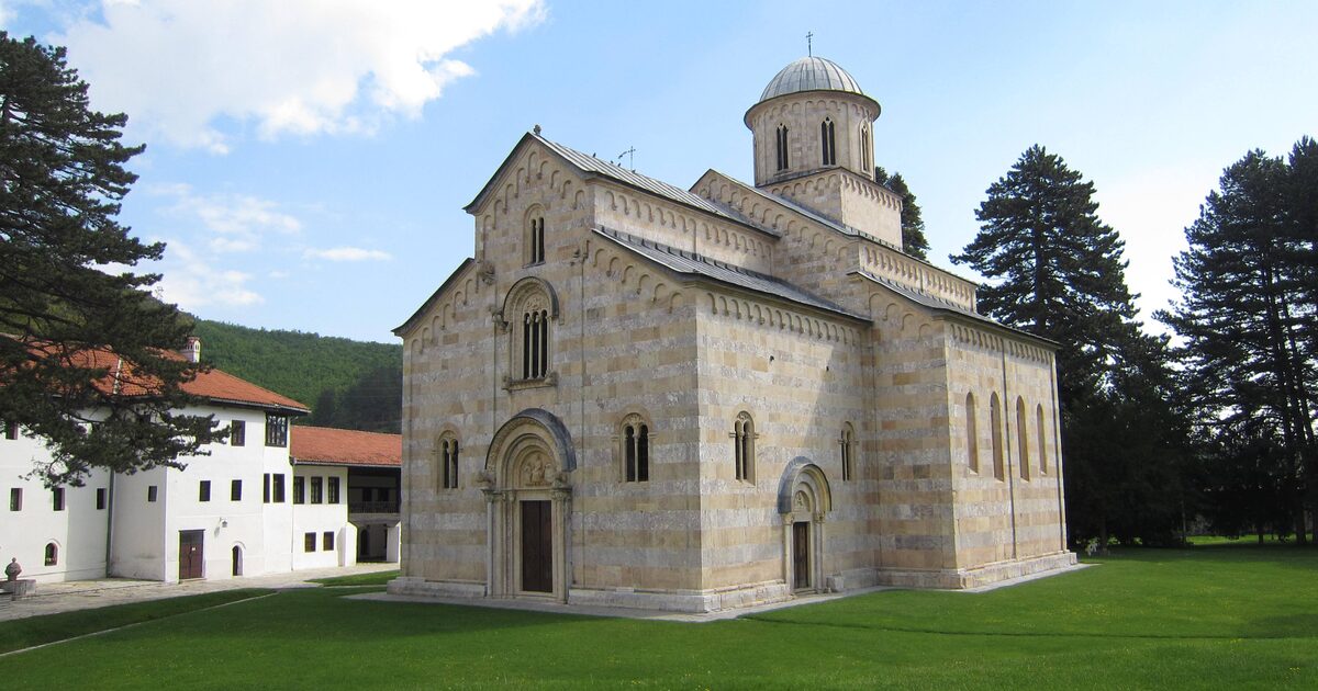 The striped marble Church of Christ Pantocrator at Visoki Dečani on its green lawn, with the white monastery lodgings, tall pines and a wooded hillside behind.